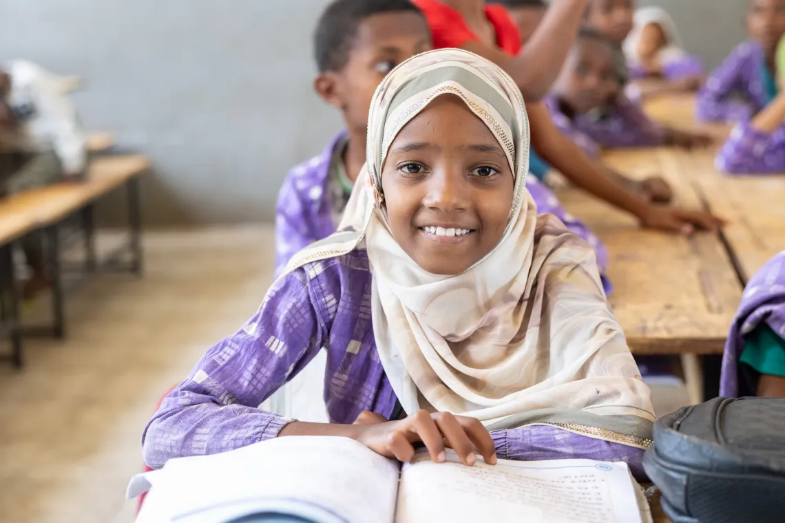 A young girl smiles to a camera siting on a desk and opening her books 