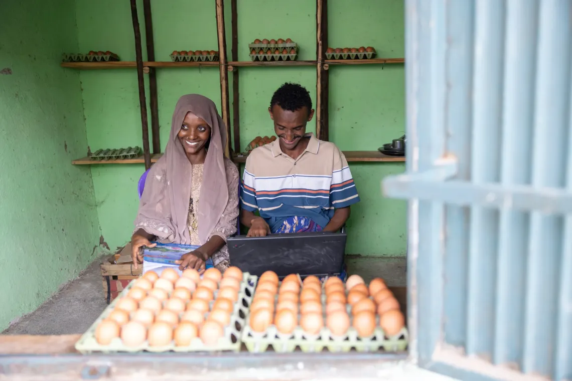 Two shopkeepers with trays of eggs use a laptop, smiling in a small green storefront.