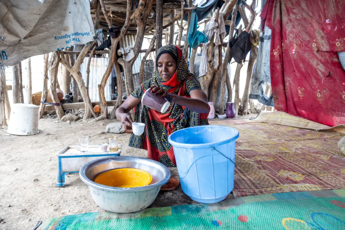 Woman washes dishes, seated on mat beside a blue bucket.