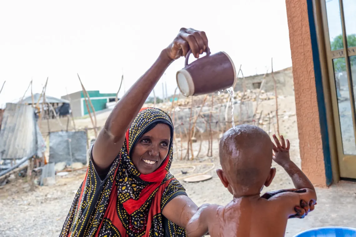 Smiling woman pours water from a mug to bathe a small child outdoors.