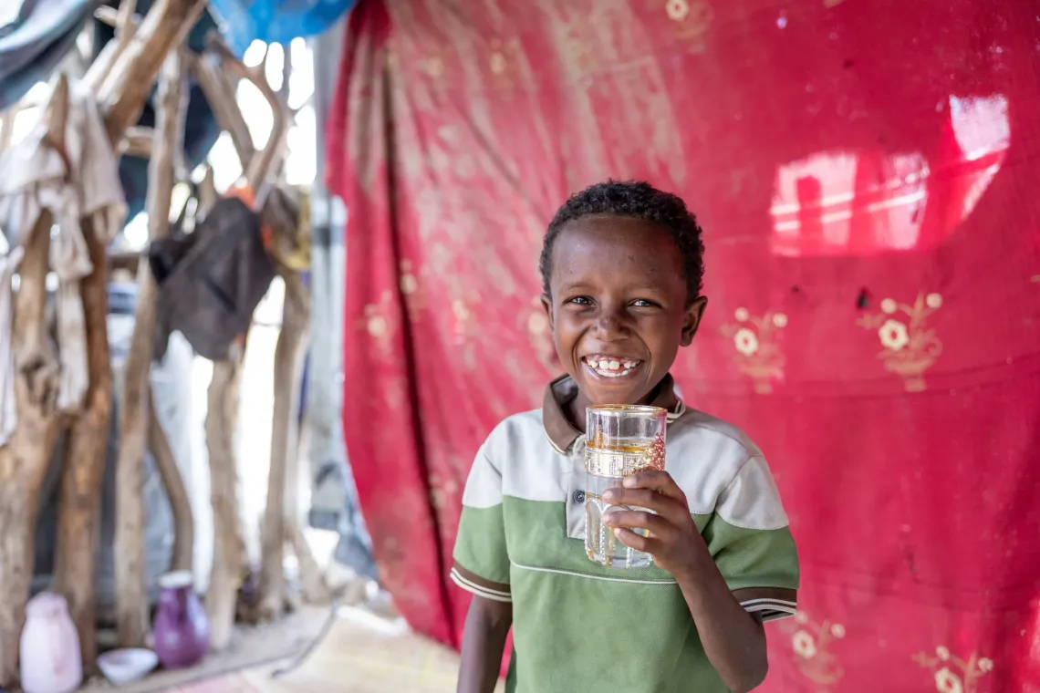 Smiling child holds a glass of water, standing in front of a red patterned cloth backdrop.