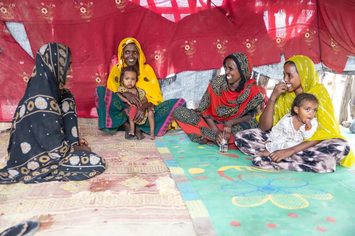 Women in bright clothing sit on floor mats inside a red tent, talking and smiling together.
