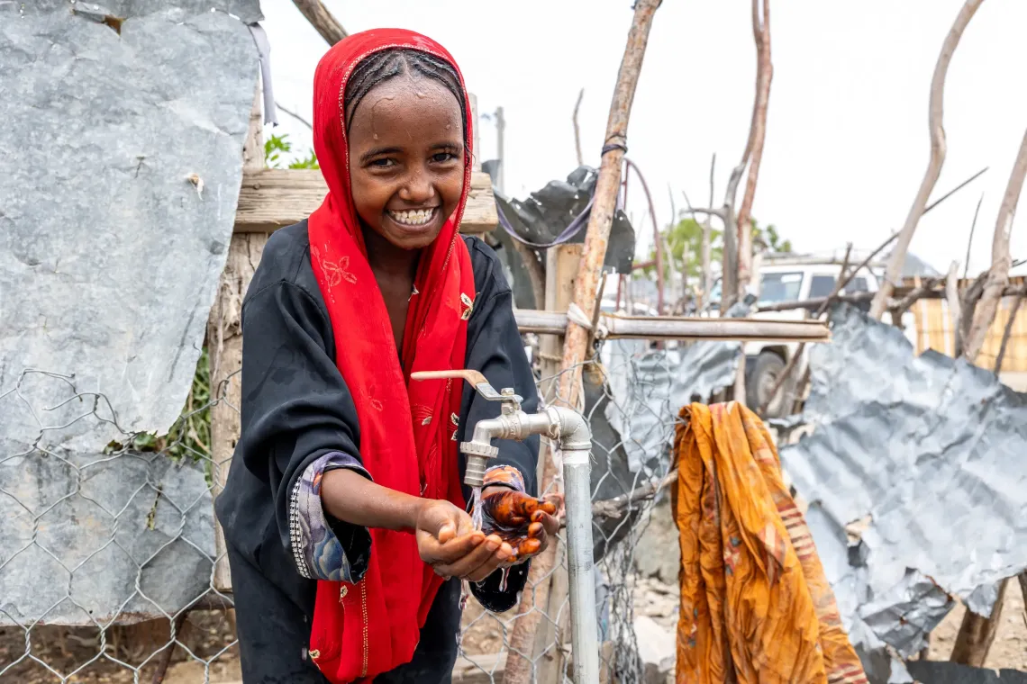 Smiling girl in red headscarf washing hands at an outdoor water tap in a rustic setting.