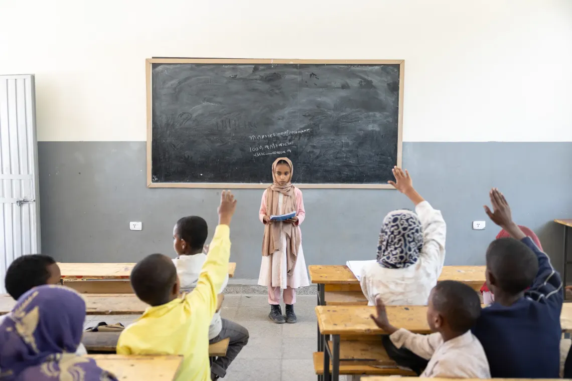 A young girl leads a question and answer session in a classroom standing in front  of her friends.    
