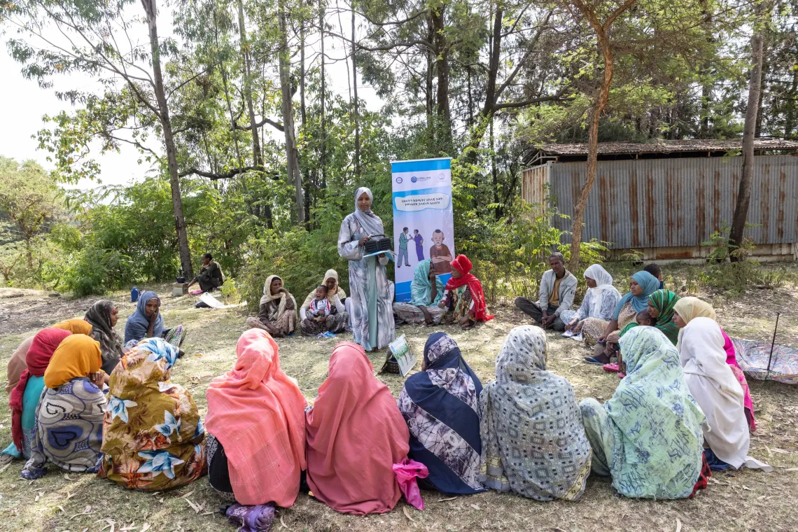 A woman teaches a lesson holding a radio while group of women sit in circle. 