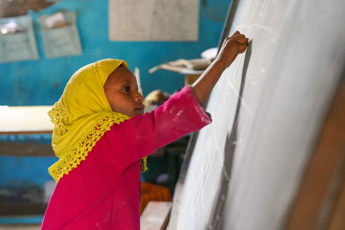 A six year old girls stretches her hand as high as possible to write on a backboard.