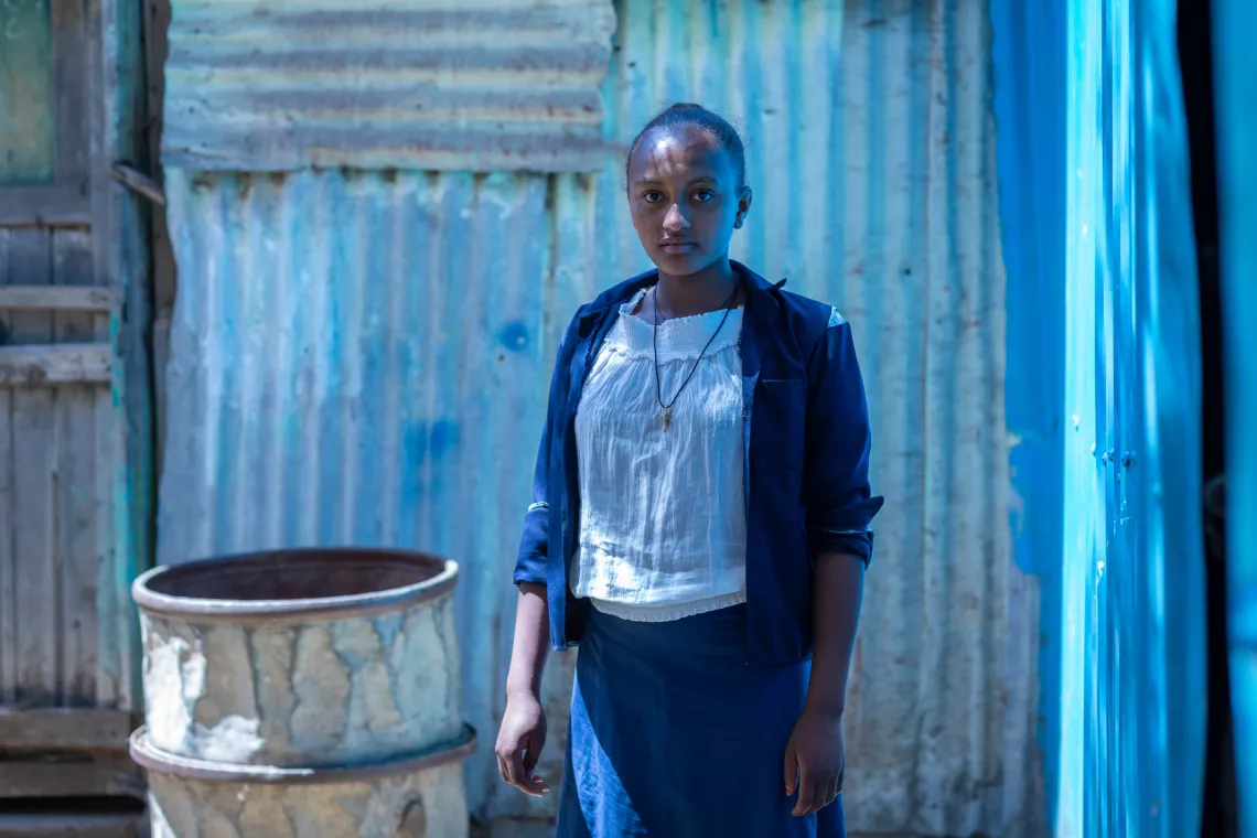A girl with a navy jacket stands outside in front of a corrugated iron wall