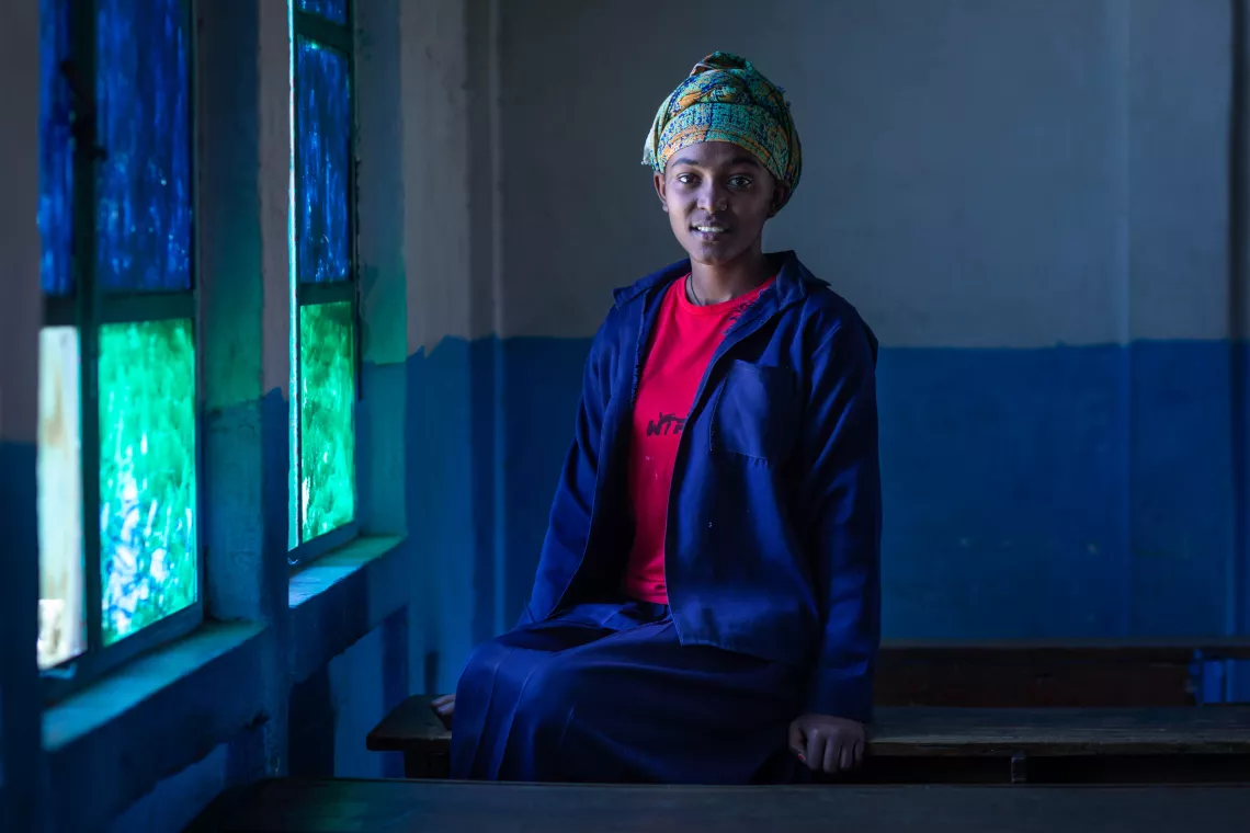 A girl with a green headscarf and a navy blue jacket sits on a classroom table. 