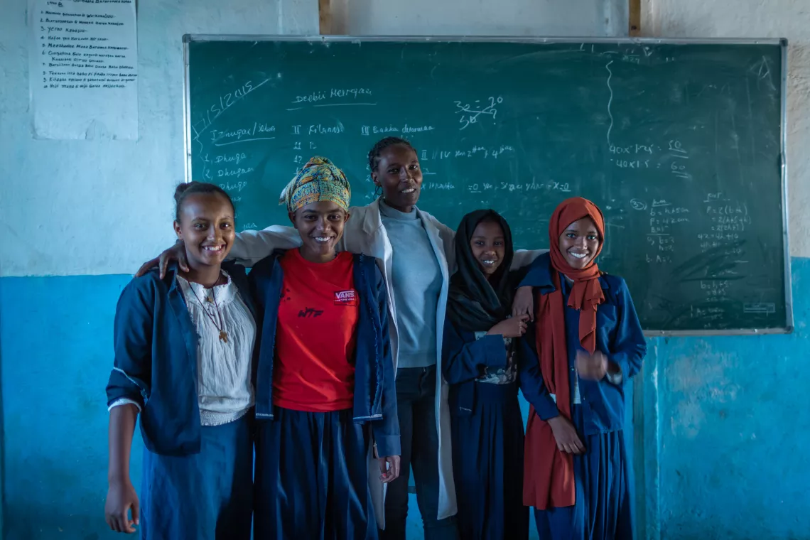 A teacher stands in front of a blackboard with two smiling teen girls on either side of her.