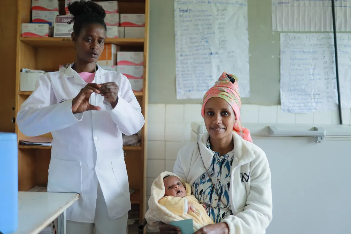 A mother holding her baby in her arm and posing the camera while a health worker is standing next to her