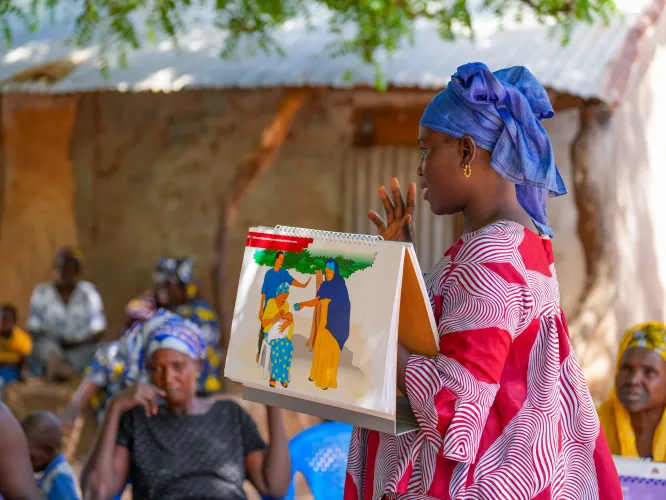 Halima Mbye leads a community session on exclusive breastfeeding in Cheken, North Bank Region. The counselling card (Health and Nutrition flipchart) reminds families to give only breast milk for the first six months.