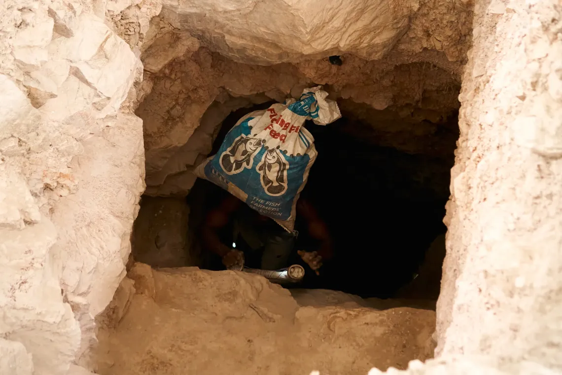 n Anfoega Adame Newtown Ghana, a miner is seen inside the kaolin tunnel carrying a bag of excavated clay on his head while holding a flashlight to guide his way. The narrow passage and rough walls show the depth and difficulty of manual extraction in the mine. 