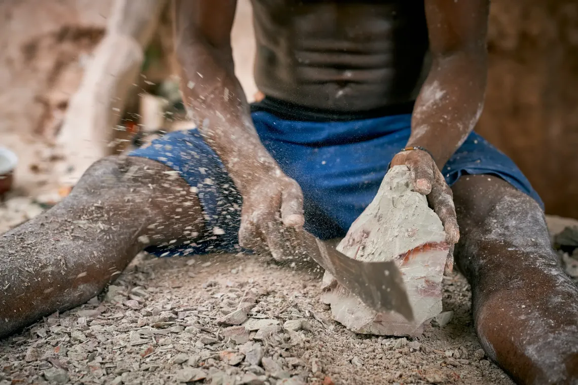 Bless, aged 33, is seen outside the kaolin mine breaking down chunks of clay to separate iron ore from the kaolin. Each precise strike of his hammer sends dust into the air, settling on the ground and bags of sorted material nearby. The scene shows the careful manual process that ensures the purity of the clay before it is collected for further use.