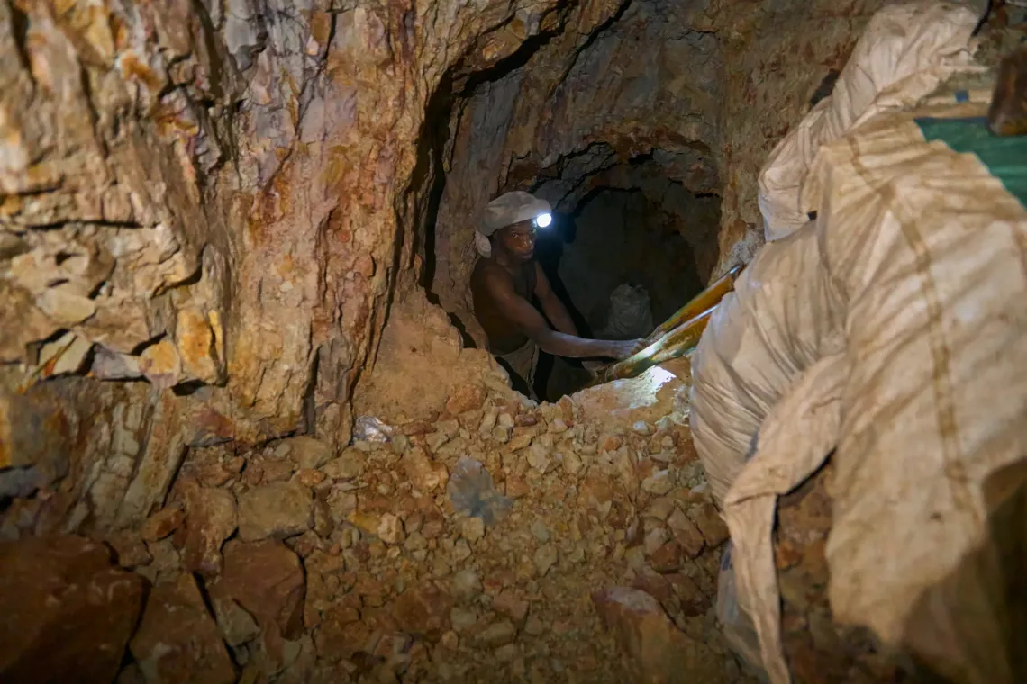 n Anfoega Adame Newtown Ghana, a miner is seen inside the kaolin tunnel carrying a bag of excavated clay on his head while holding a flashlight to guide his way. The narrow passage and rough walls show the depth and difficulty of manual extraction in the mine. His stance and grip reflect the strength and balance required to move through the confined space.  This image captures the physical effort and focus of underground labor, illustrating the determination and skill behind Ayilor&rsquo;s production in Ghana&rsquo;s k