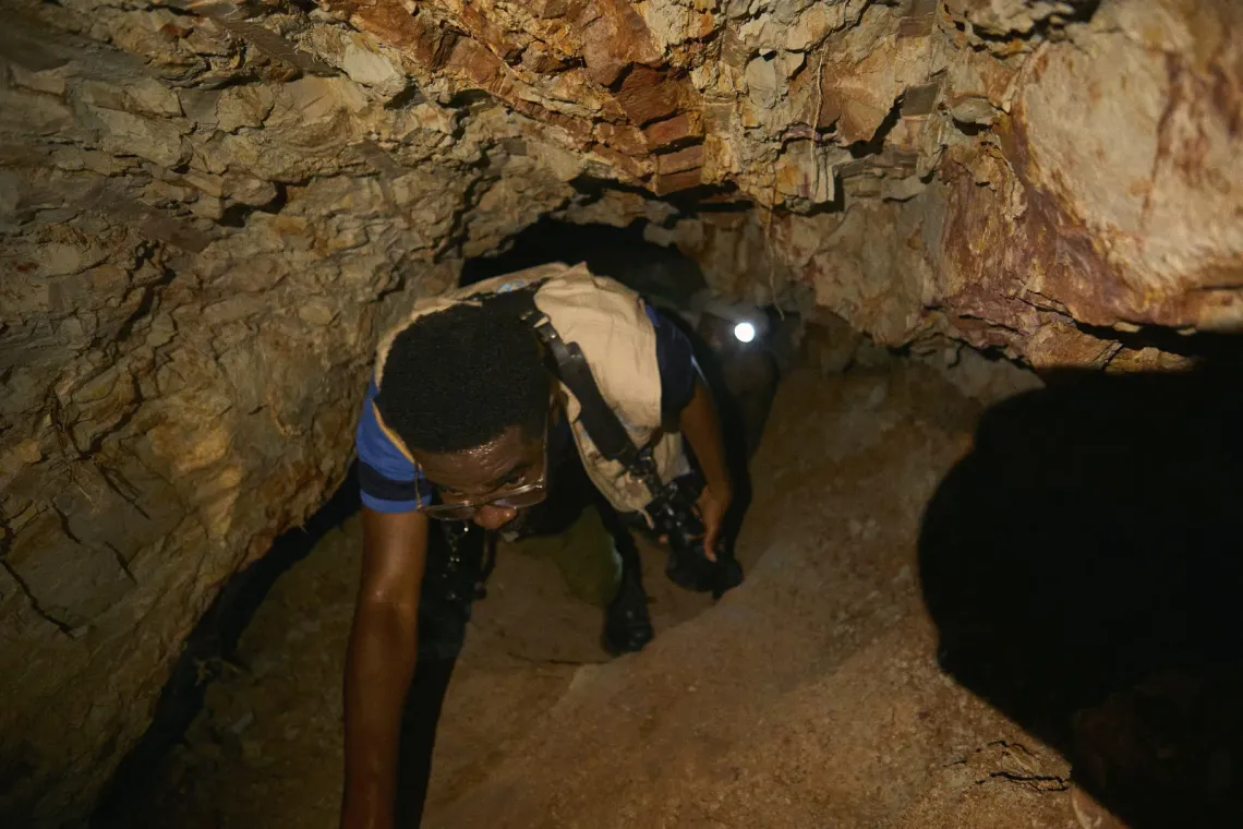 UNICEF consultant Eric crawls carefully through the narrow passage of the kaolin mine, his camera strapped to his chest as he documents the conditions below. Behind him is Francis Dompeh, the 37‑year‑old miner guiding the way with his headlamp casting a steady beam through the dark. The rock walls press close on both sides, their layers revealing the geological history of the region and the depth of manual labor required to extract the clay.