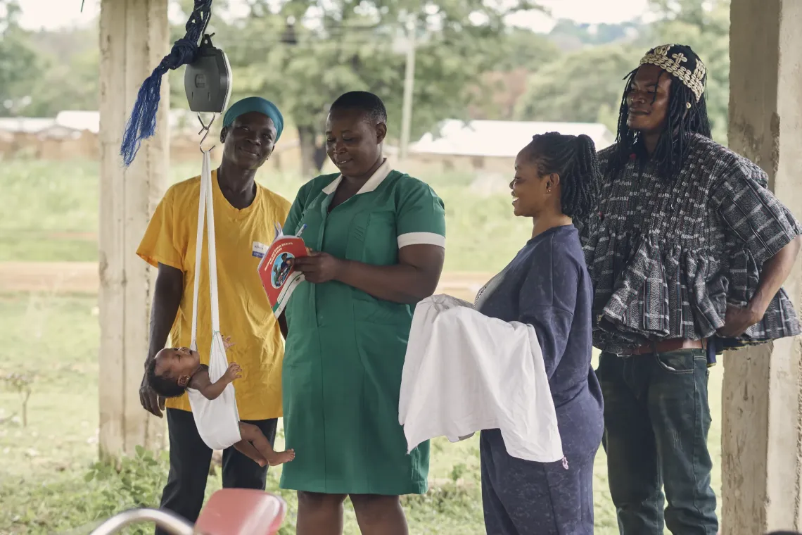 At the Gundoug/Mihiiboug Community-based Health Planning and Services (CHPS) compound in Ghana's Upper East Region, a nurse carefully weighs a baby using a hanging scale while community health volunteer Aida Bugre (34) smiles and observes.