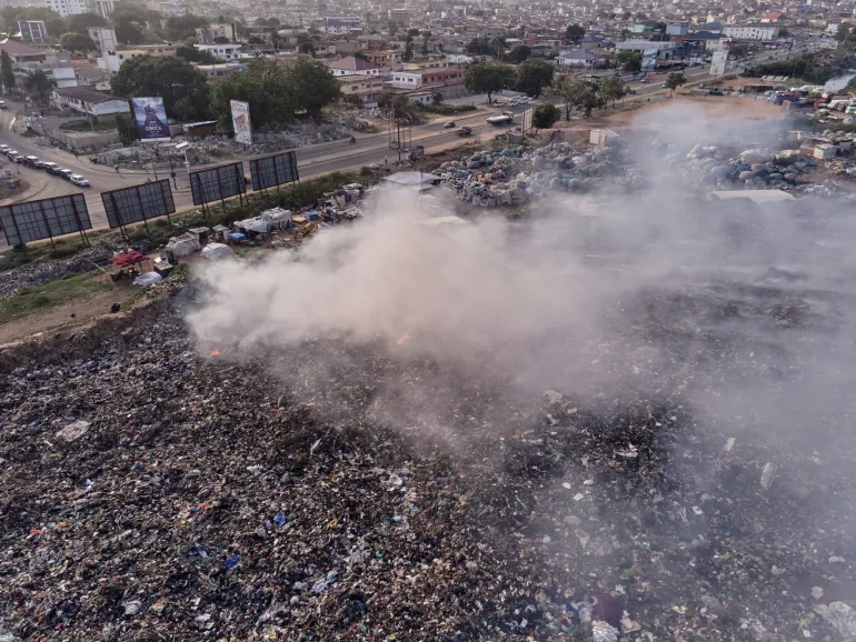 Heaps of plastic and discarded materials stretch across the landscape, with smoke rising from open burning. 