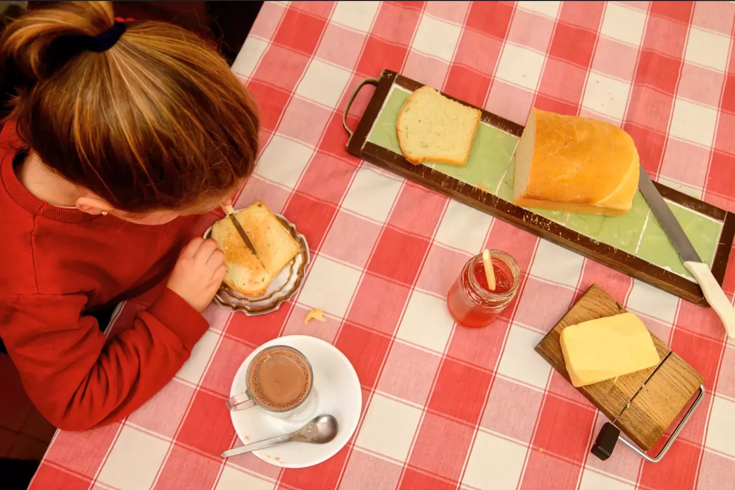 Girl child cutting a sandwich with a knife with bread and jam being set on the table.