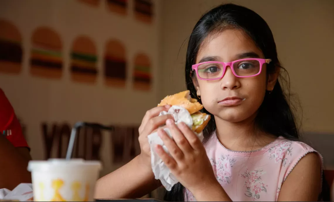 Brunette girl with pink eye glasses eating a burger straining directly at the camera