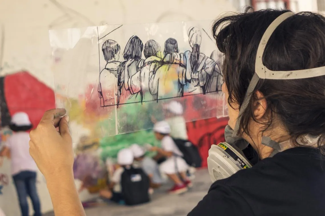 A woman with a painting respirator examines an image drawn on transparent plastic against a mural behind it