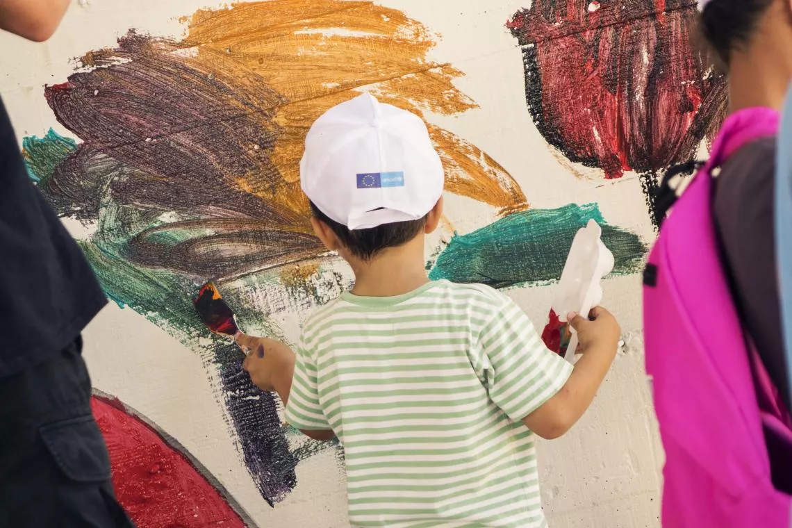 A young child wearing a white cap with a European Union logo on it holds a paint tray and paintbrush paints on a wall