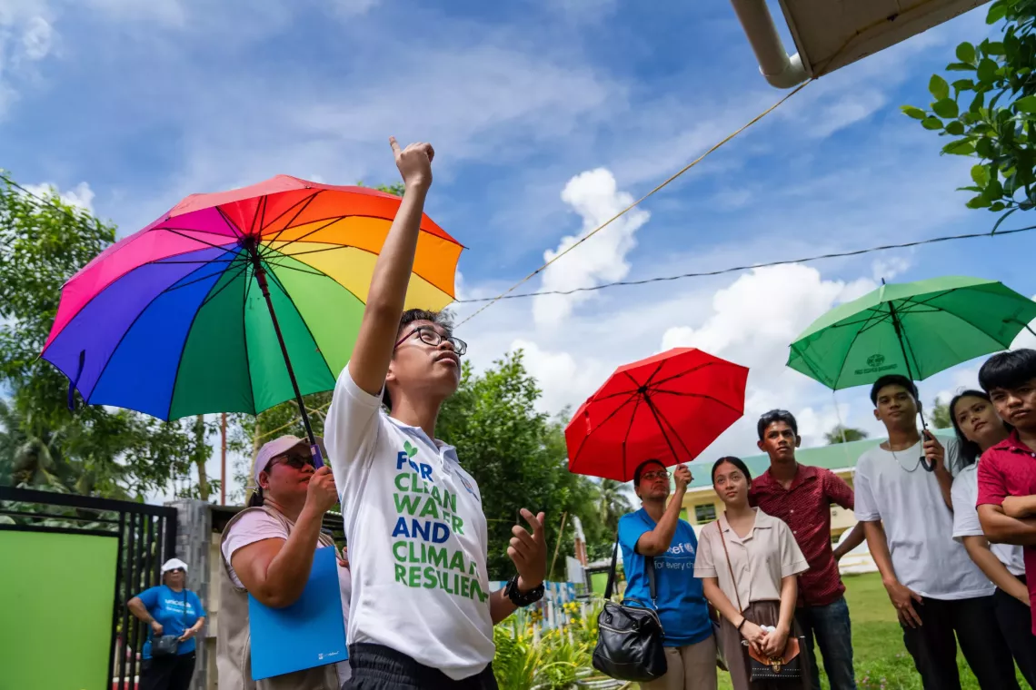 High school students under colorful umbrellas watching a science demonstration on water filteration