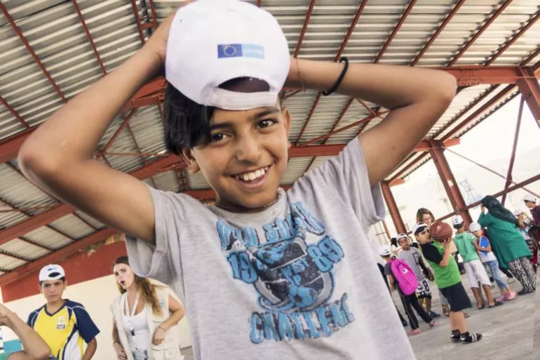 Child in the foreground putting on a white cap with an EU logo on it with other children and adults standing in the background