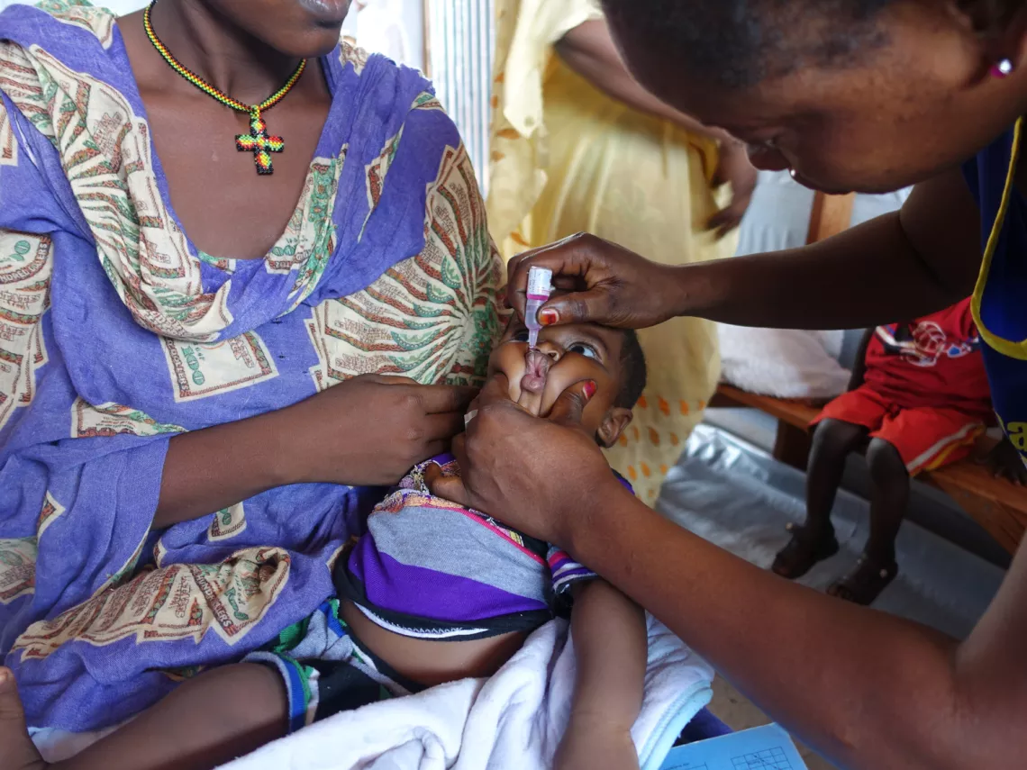 A baby being held by his mother while a nurse leans over with an oral vaccination bottle.