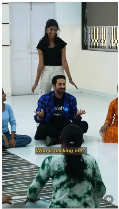 UNICEF India National Ambassador Ayushmann Khurrana sitting cross-legged on the floor with a group of young people in a community centre, with the subtitle "Who is hacking me?" suggesting a discussion on online safety.