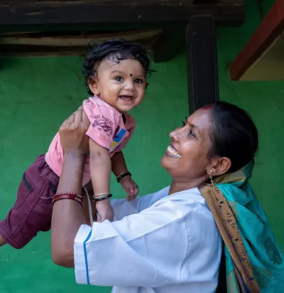 A joyful Indian woman in a white uniform lifts a giggling baby high in the air outside a green-walled rural home, both beaming with delight.