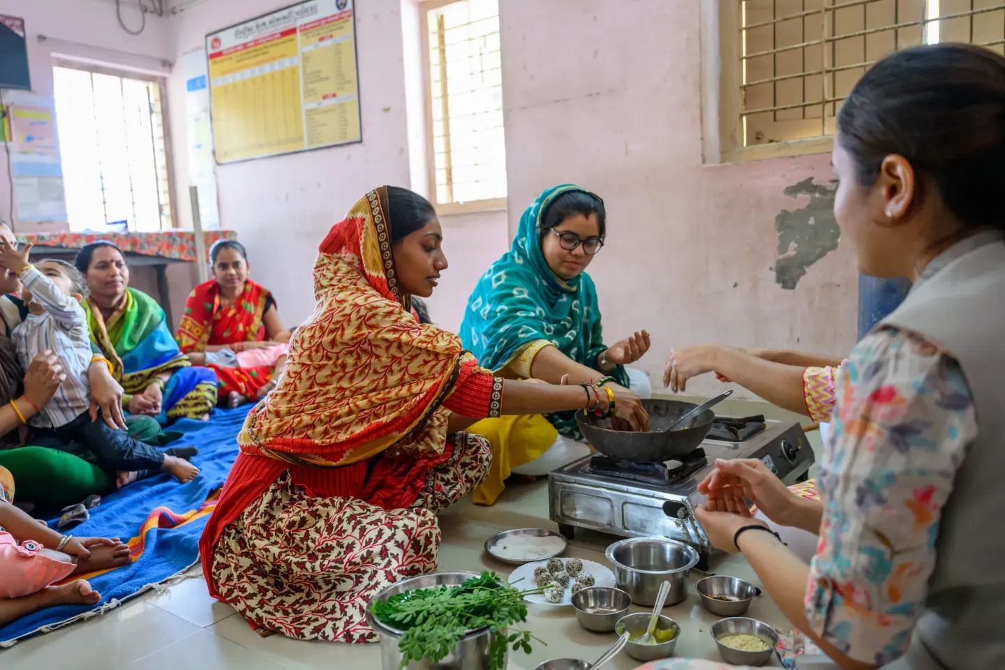 Women participate in a nutrition cooking demonstration using local ingredients including moringa leaves and energy balls at a community centre, watched by other women and children seated on mats.