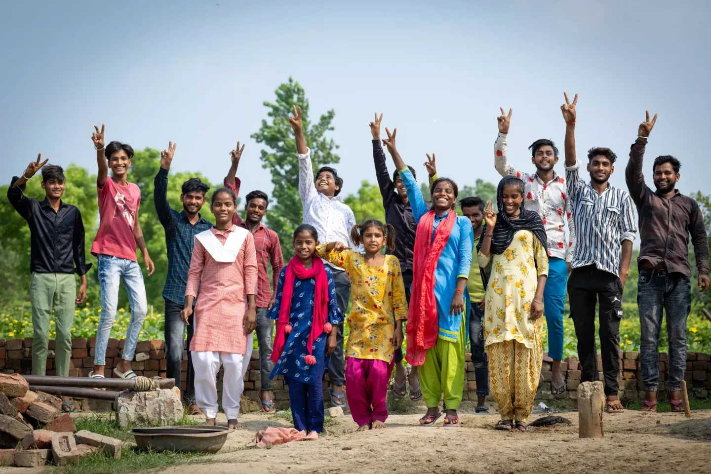 A large group of joyful Indian adolescents in colourful clothing raise peace signs and cheer together outdoors in a sunny rural village setting.