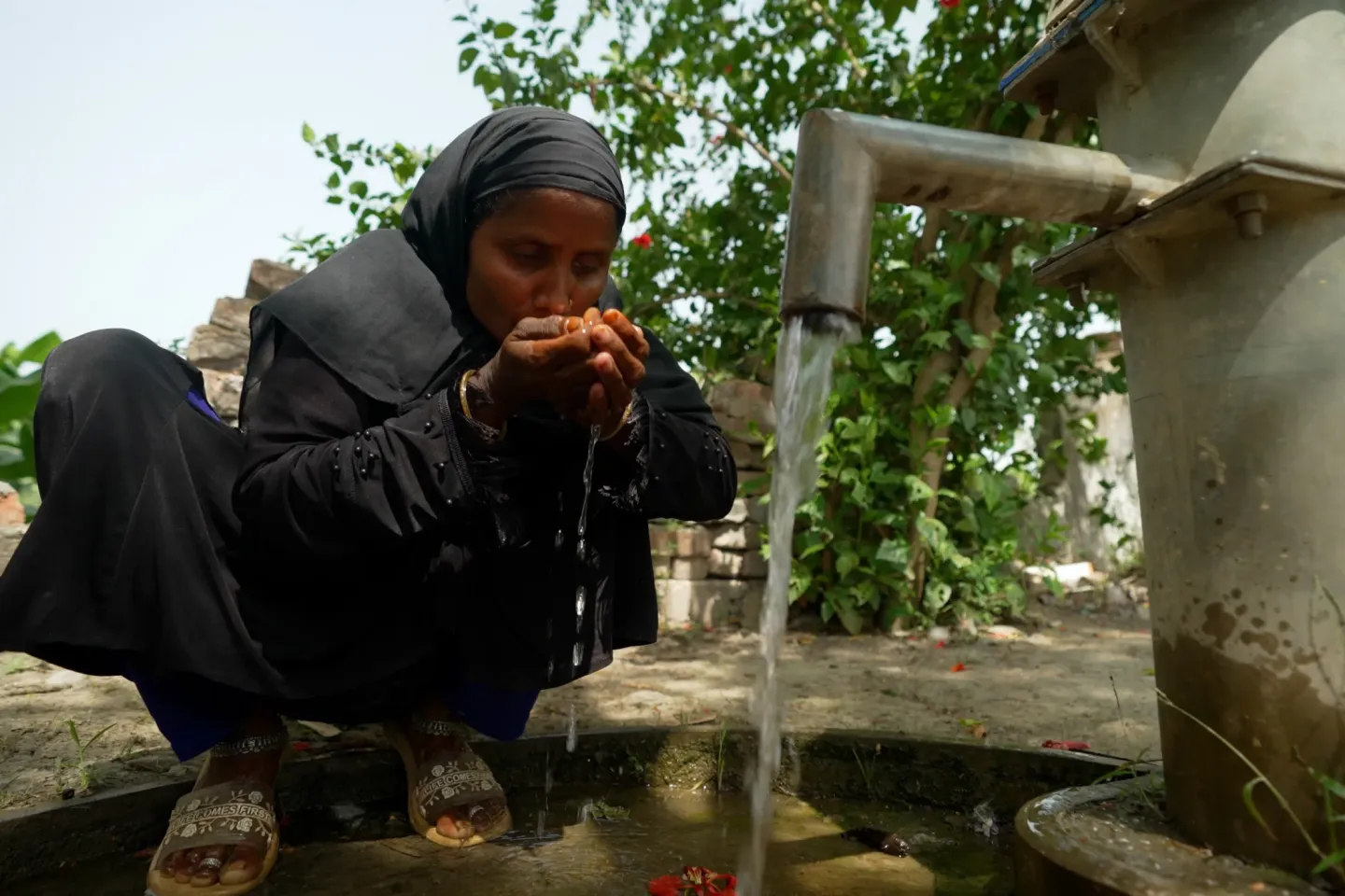 A woman in a black hijab crouches beside a community water tap, cupping her hands to drink clean running water in a rural village setting.