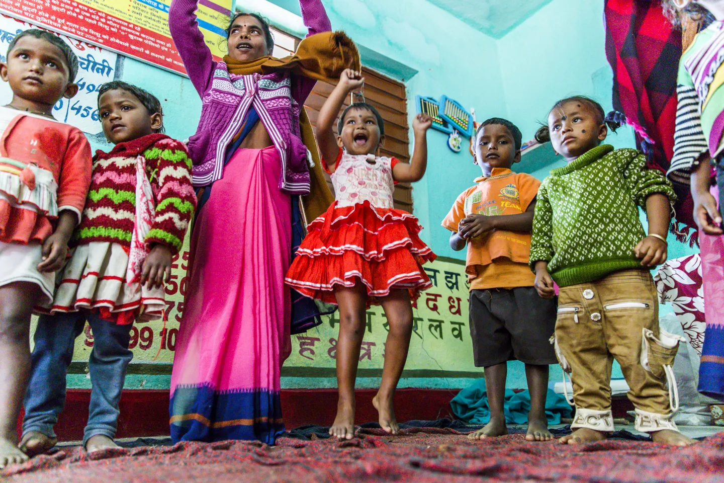 A group of young Indian children play an energetic indoor game barefoot in a colourful classroom, with some running and others cheering from the sidelines.