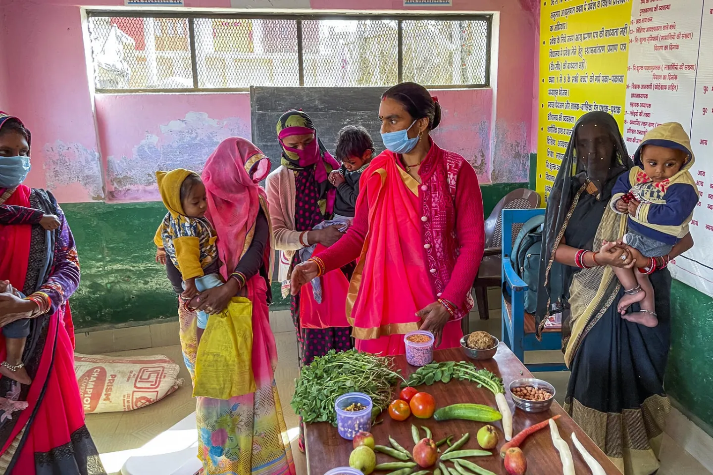 A frontline health worker in a pink saree and face mask conducts a nutrition demonstration for mothers with young children at an Anganwadi centre, with fresh vegetables and foods displayed on a table.