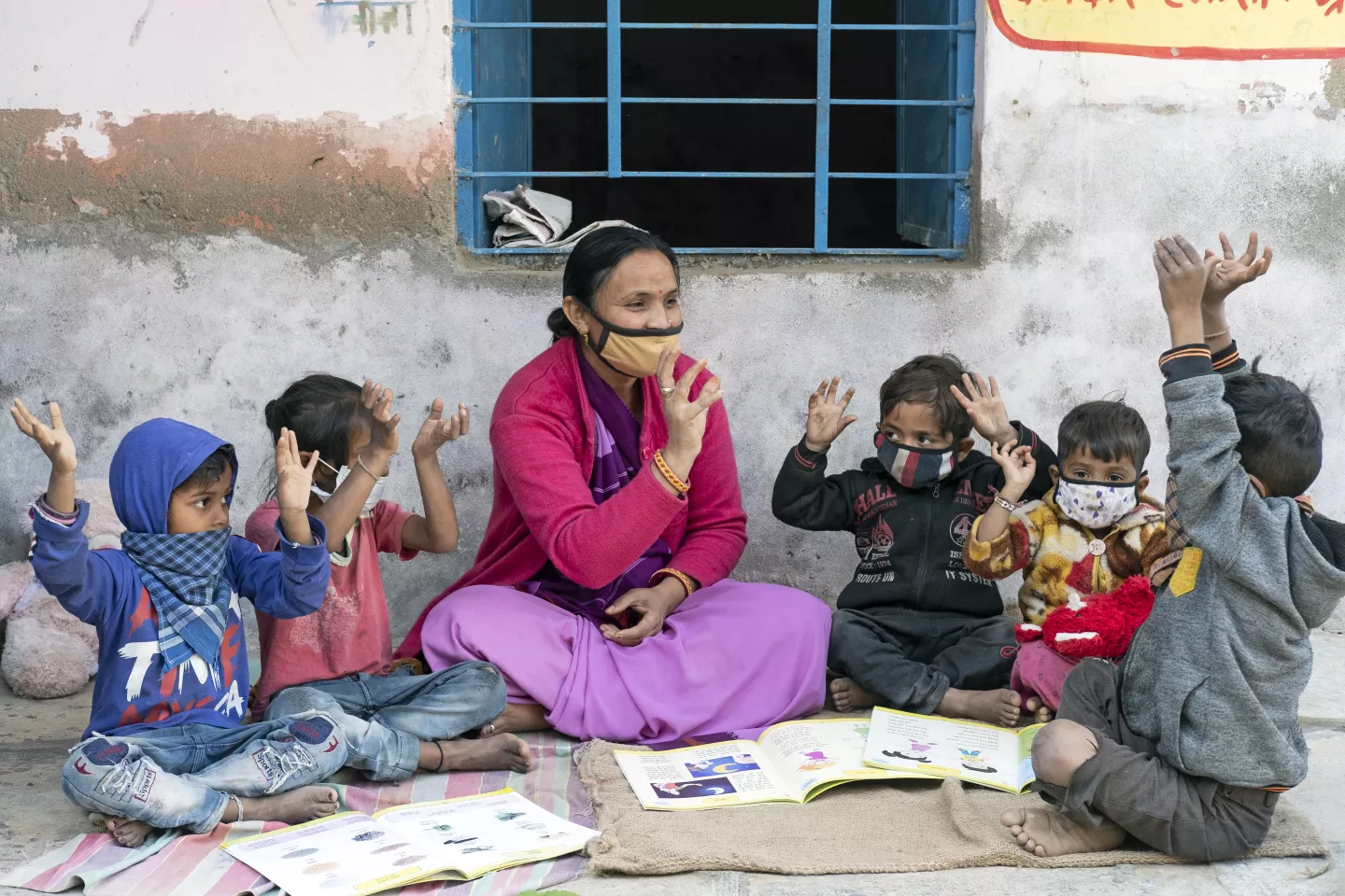 An Anganwadi worker in a pink saree and face mask conducts an outdoor early learning session with five young children, all raising their hands and following along with colourful picture books open in front of them.