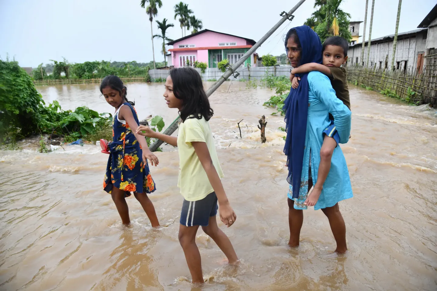 A woman carries a young boy on her back as two girls wade beside her through muddy floodwater in a village in Assam, India.