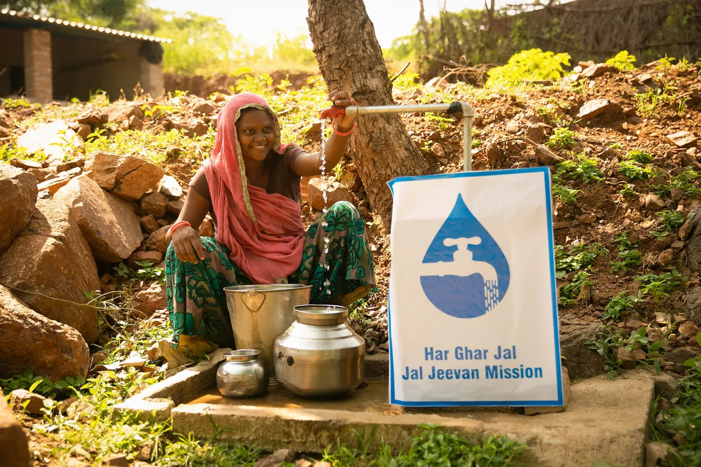 A smiling woman in a pink dupatta turns on a community water tap beside a 'Har Ghar Jal &mdash; Jal Jeevan Mission' sign, filling steel vessels in a rural setting.