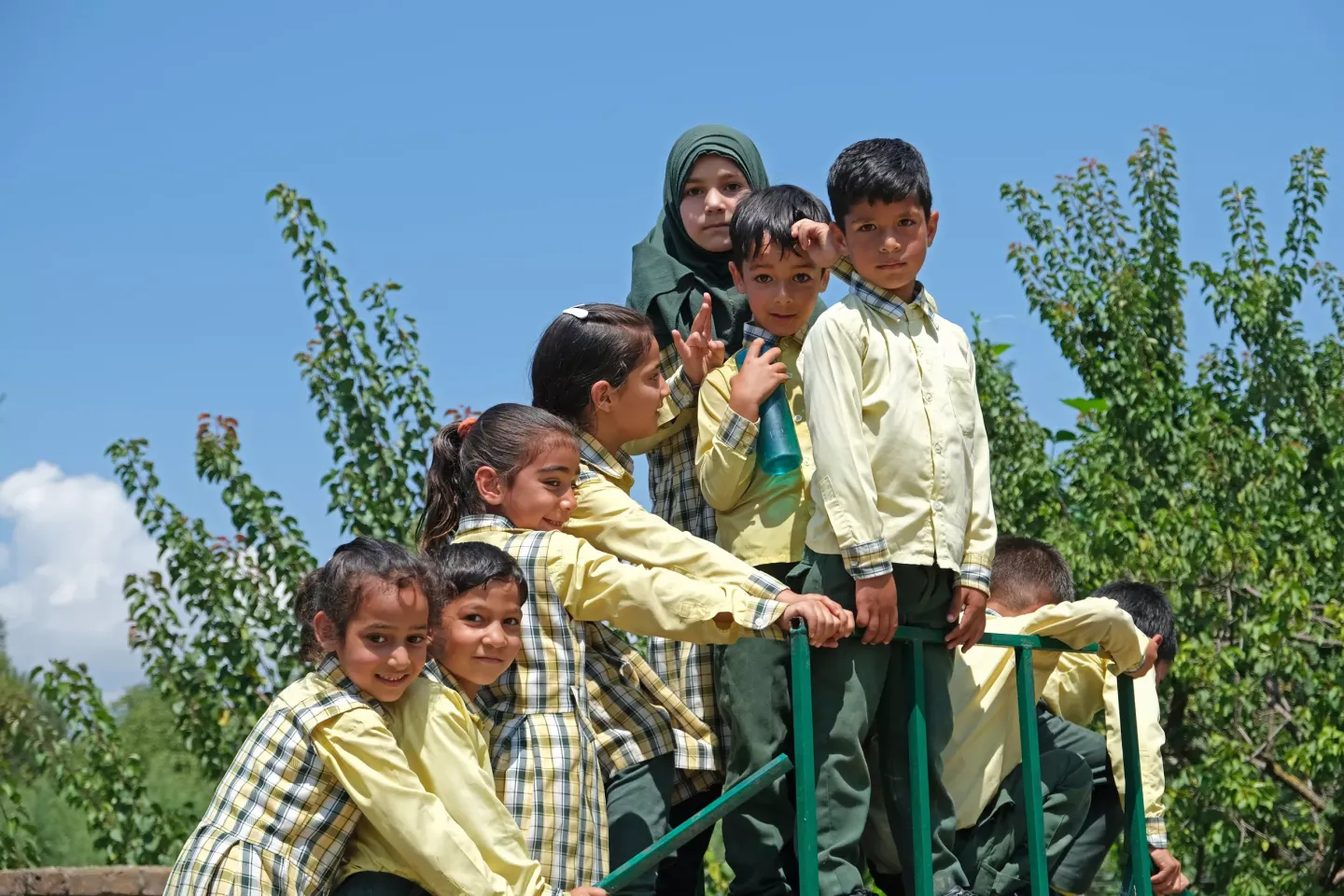 A group of young schoolchildren in yellow checked uniforms cluster together on outdoor playground equipment under a bright blue sky, with one girl wearing a green hijab standing behind them.