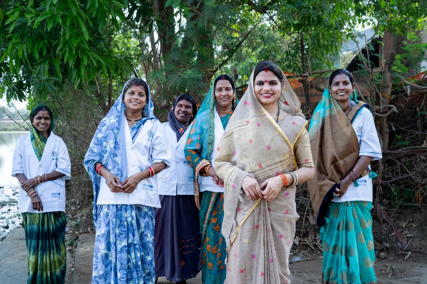 A group of six smiling Indian women &mdash; some wearing UNICEF "for every child" white coats &mdash; stand together outdoors near a river, representing frontline health workers and community leaders.