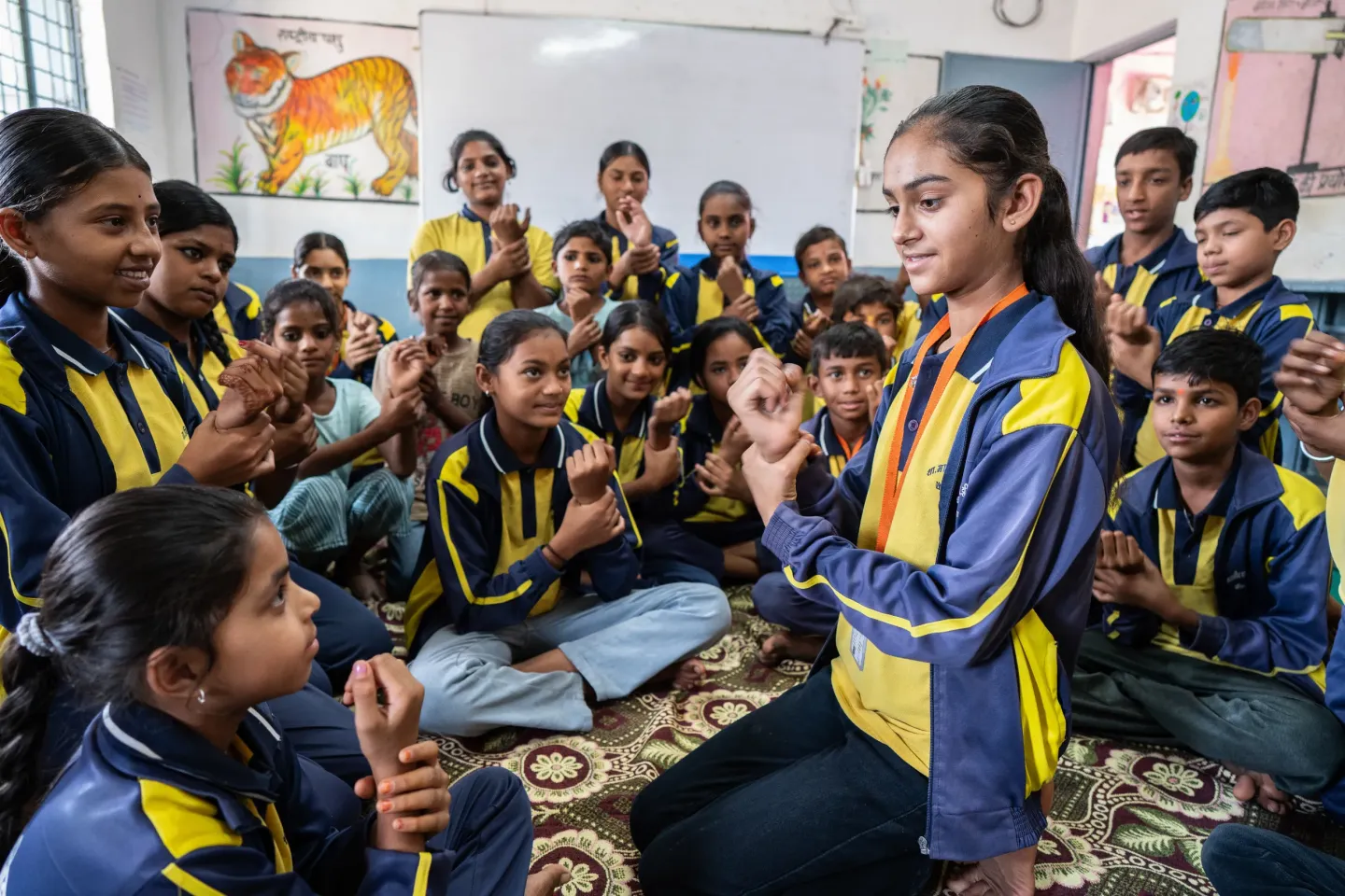 Rakshita Yadav, 13, demonstrates proper handwashing technique to her classmates seated on the floor of a school classroom, all imitating her hand movements."