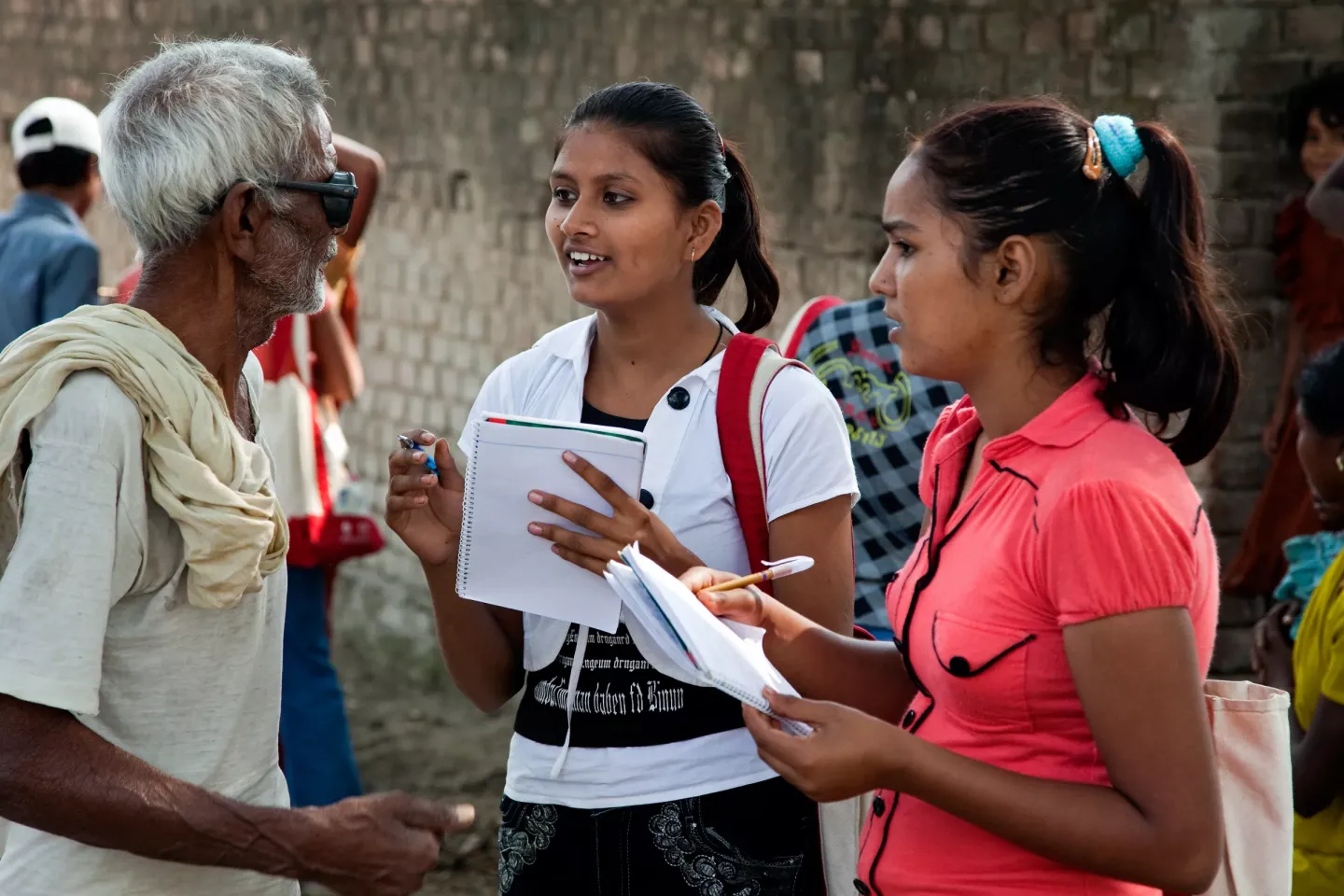 Two young Indian women with notebooks and pens interview an elderly man on a busy street, in what appears to be a community outreach or field survey.