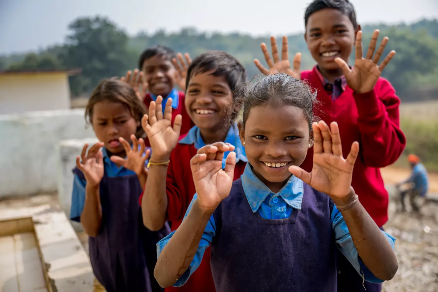 A group of joyful Indian schoolchildren in blue and red uniforms show their clean hands to the camera, smiling broadly outdoors.