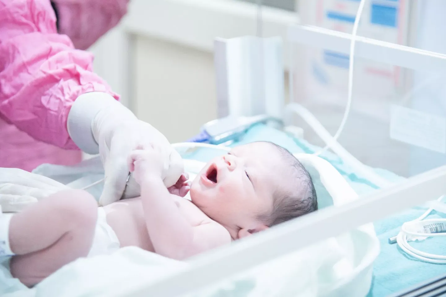 A newborn baby lies in a hospital bassinet, mouth open and eyes partly closed, as a gloved healthcare worker gently holds the infant's hand.