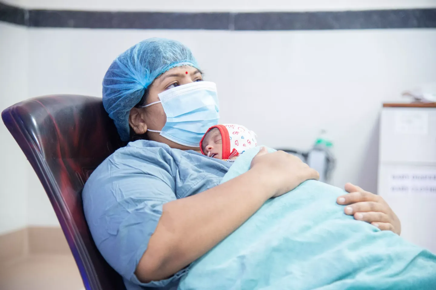 A new mother in a hospital gown, surgical cap, and face mask holds her sleeping newborn close to her chest in a maternity ward, practicing skin-to-skin care.