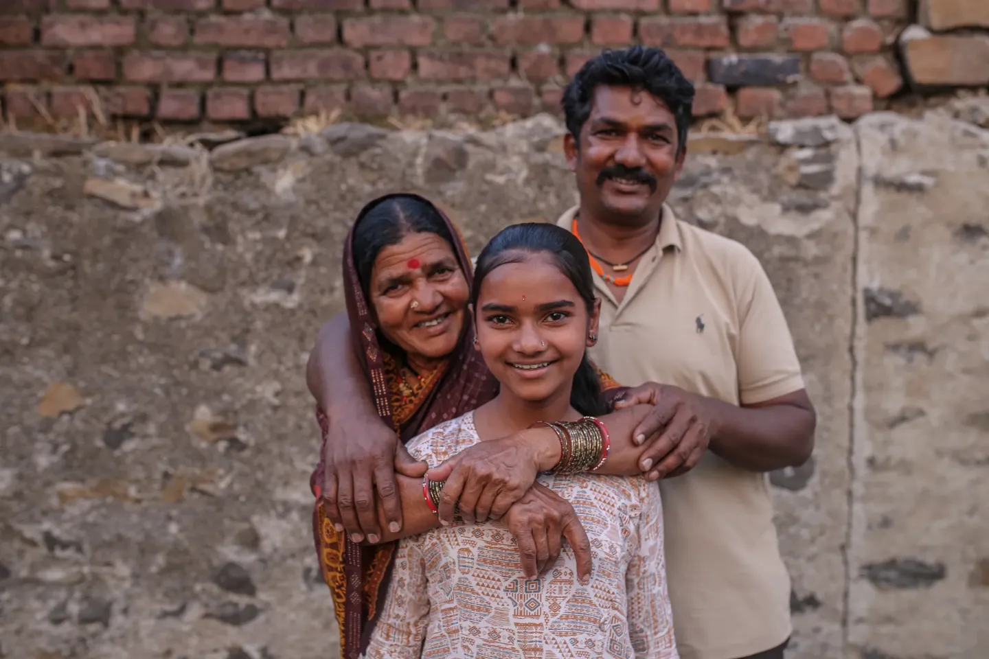 A smiling Indian girl, warmly embraced by her grandmother in a saree and her father, poses together outside their home in a rural village setting.
