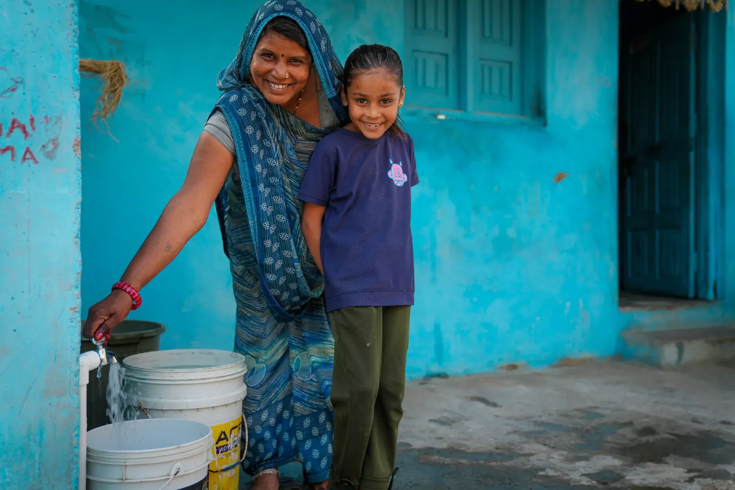 Smiling woman in a sari and young girl stand by white buckets against a bright turquoise wall.
