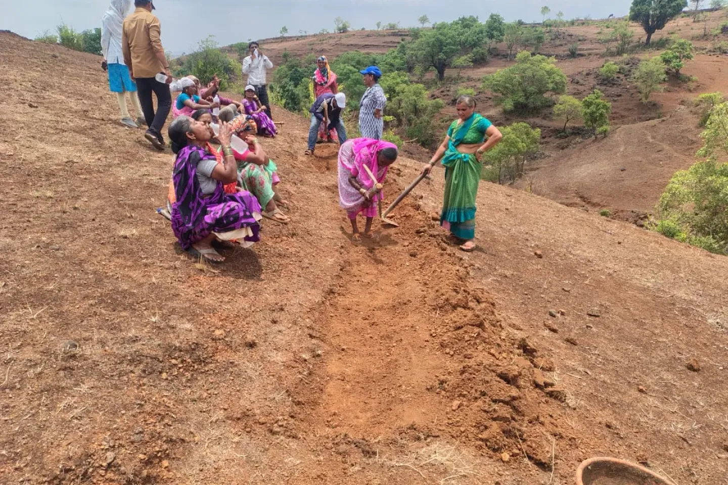 A group of rural women in colourful sarees dig and hoe a Continuous Contour Trench on a dry red-soil hillside in Maharashtra during a community Shramdaan water conservation campaign.