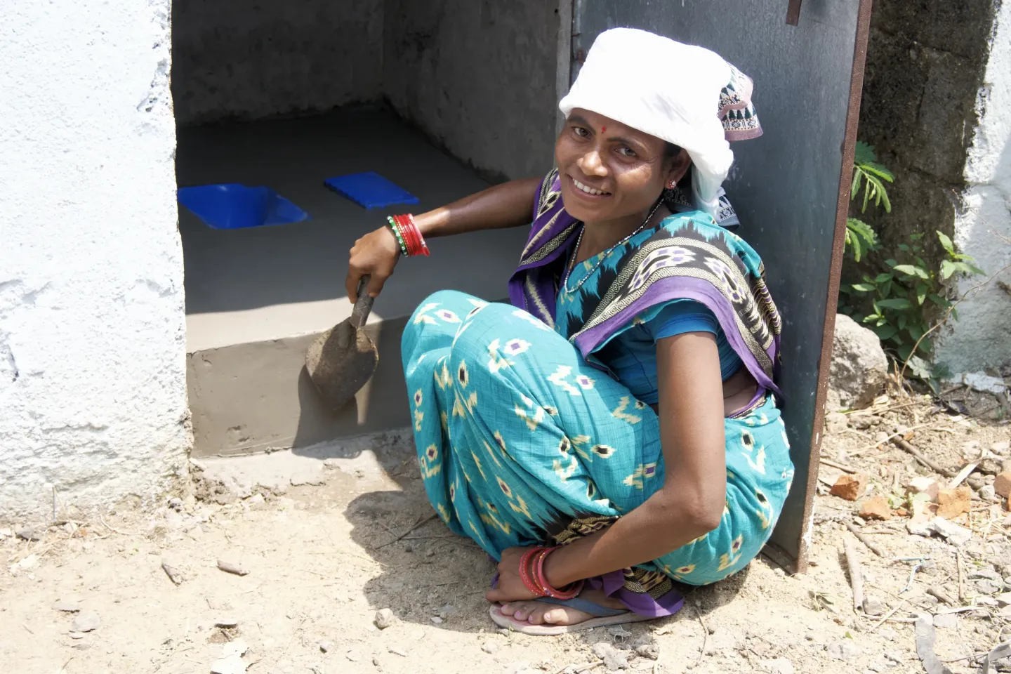 A smiling woman mason in a teal ikat saree and white head covering sits at the doorway of a newly plastered toilet she is constructing in Odisha, holding a masonry trowel.
