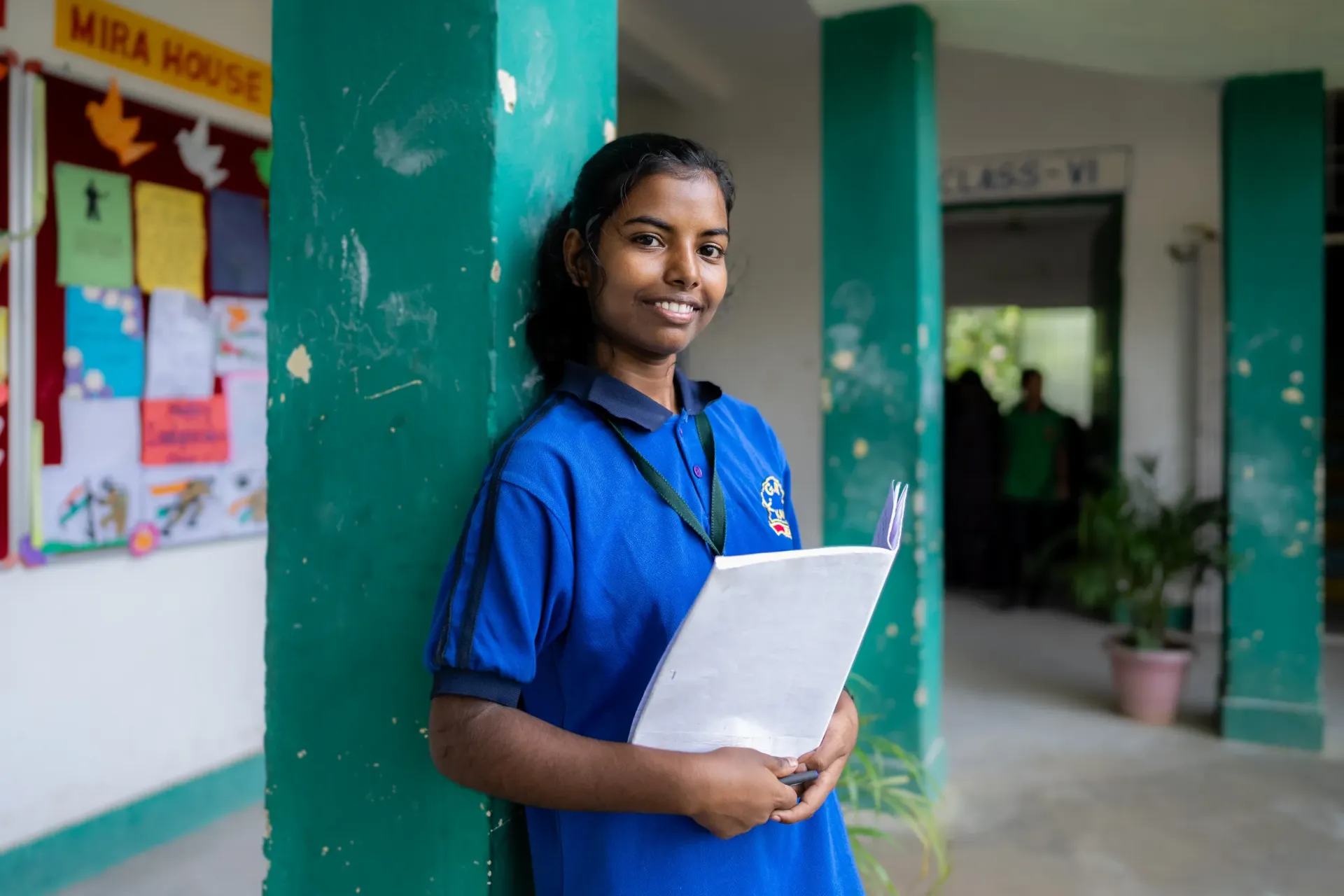 A smiling Indian teenage girl in a blue school uniform leans against a green pillar in a school corridor, holding a notebook and pen.